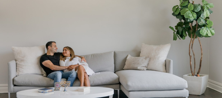 Ein Paar sitzt im Wohnzimmer auf einer hellen Couch und lächelt sich an.