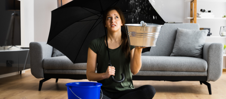 Was tun bei einem Wasserschaden? © MyPlace SelfStorage Eine Frau sitzt im Schneidersitz auf dem Boden in ihrem Wohnzimmer, mit einem Regenschirm in der Hand und einem Eimer neben sich stehen. Sie sieht traurig aus und es tropft über ihr von der Decke..