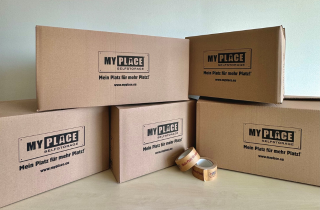5 verschieden große Kartons sind gestapelt, auf ihnen ist groß das Logo von MyPlace-SelfStorage zu sehen, davor liegen zwei Rollen Klebeband