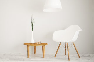 Ein weißer Stuhl steht neben einem kleinen Holztisch. Auf diesem steht eine weiße, dünne Vase mit grünen Trockenblumen