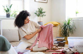 Eine Frau  sitzt in ihrem Wohnzimmer auf dem Boden und hält lächelnd einen rosa Pullover nach oben. 